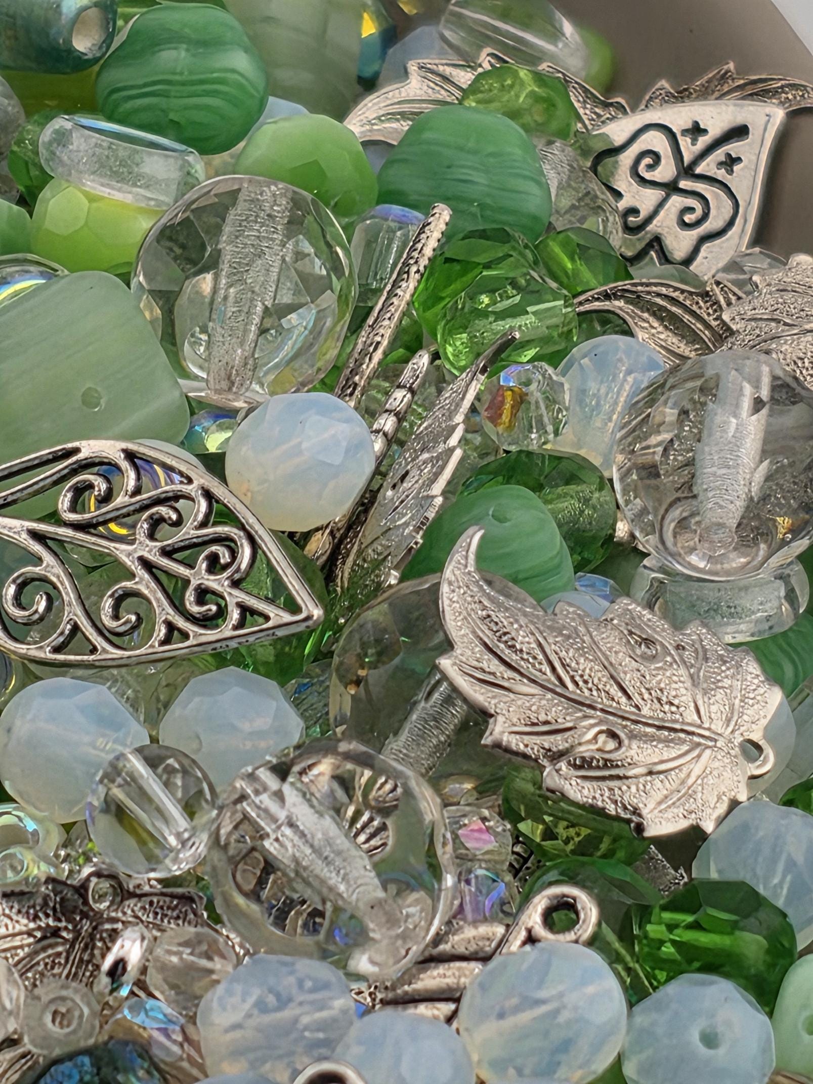 The image is a close-up of various beads, charms, and metal pieces, including clear and green glass beads, metal leaf-shaped charms, and other decorative elements.