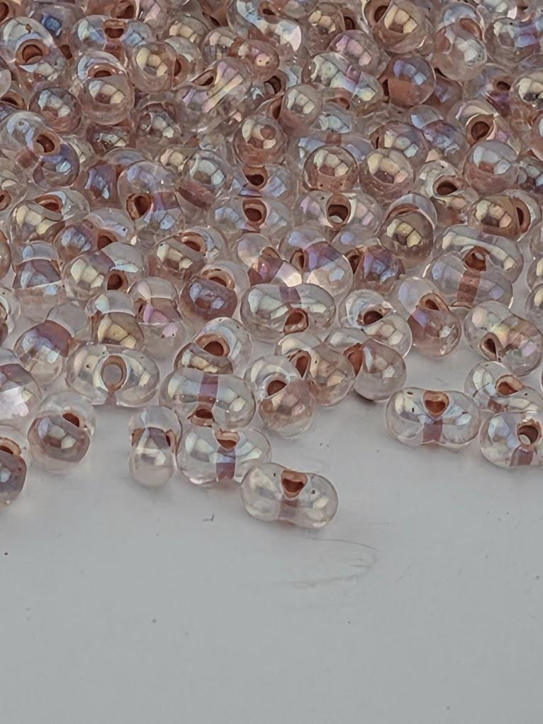 a close-up view of numerous small, clear beads scattered across a dark surface. The beads appear to be made of glass and have a transparent, iridescent quality, reflecting light in various colors. The image does not contain any text or specific details about the beads, and it does not depict any actions or movements. The focus is on the individual beads and their arrangement on the surface.