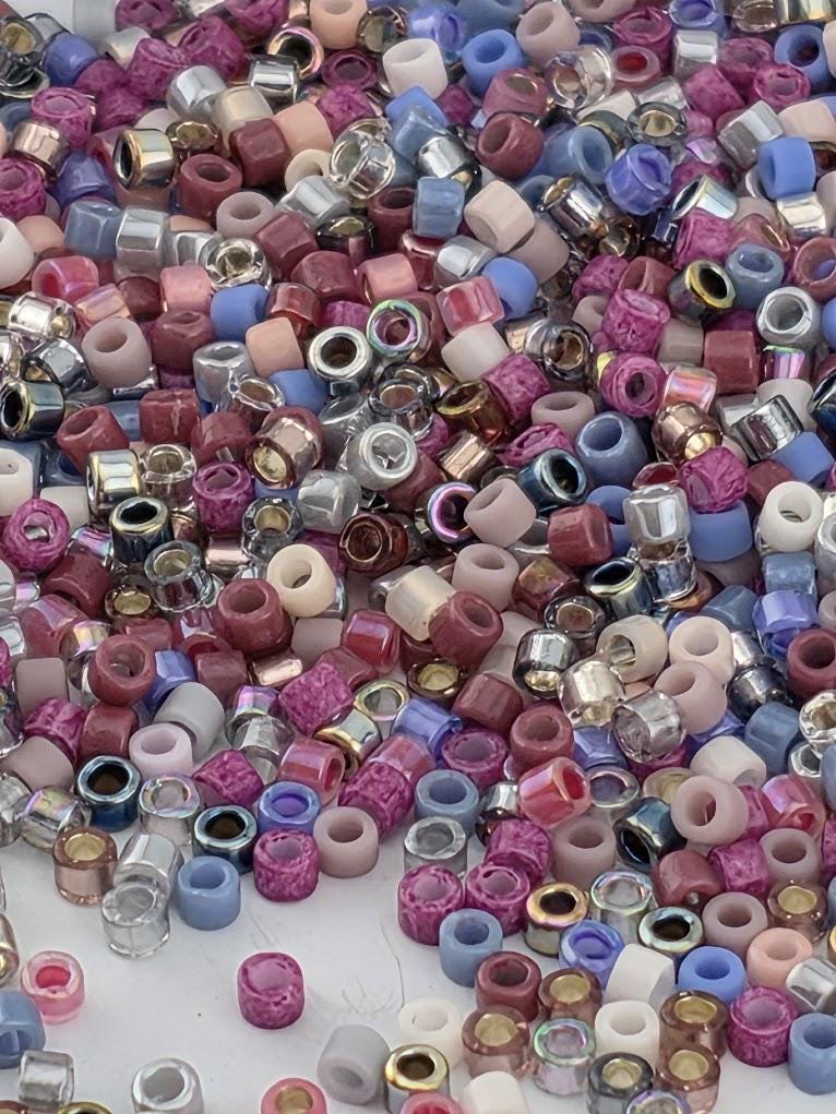 The image is a colorful assortment of various beads and small objects, including metallic and glass beads in shades of pink, purple, blue, and silver.