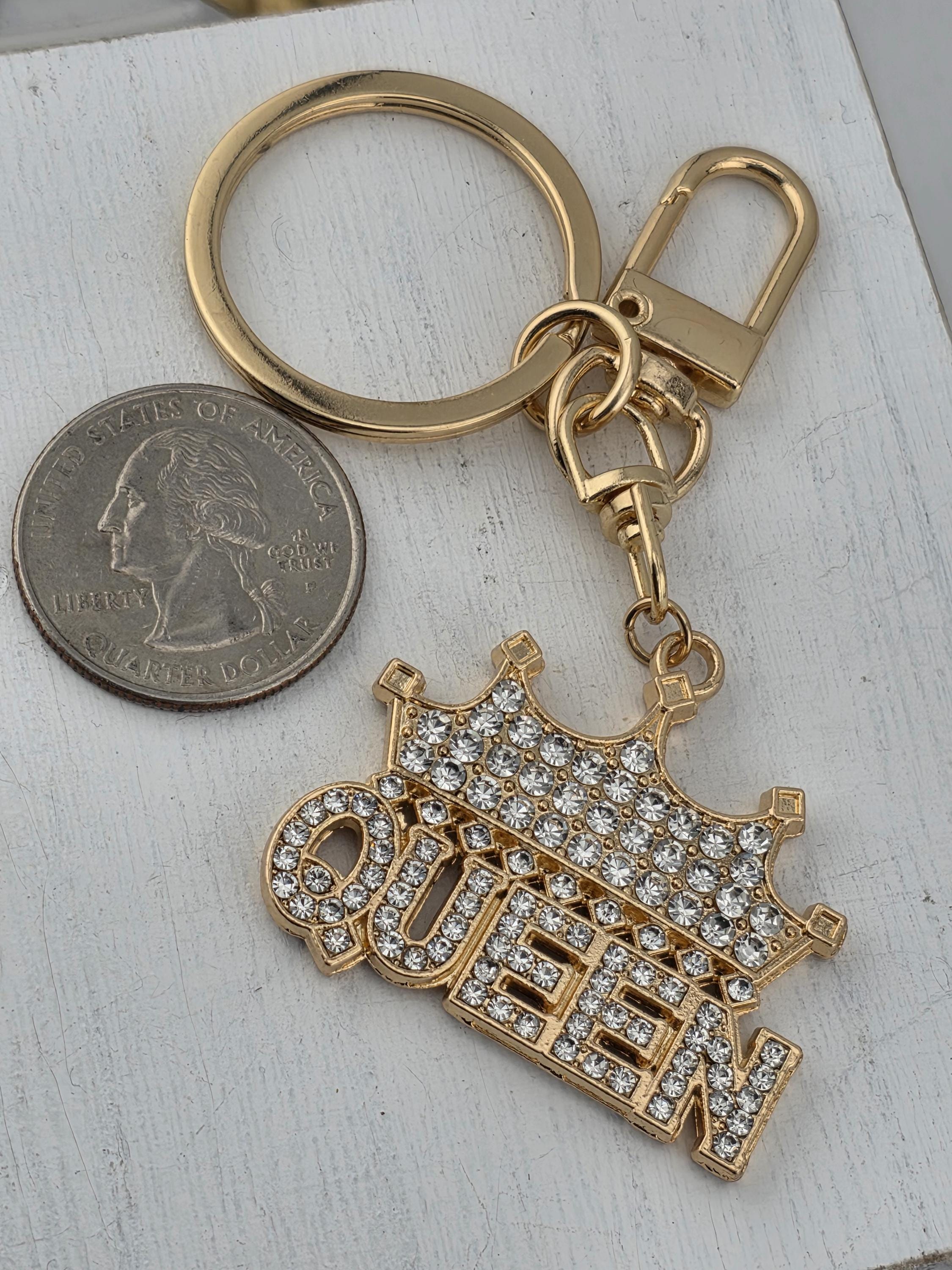 A keychain with the word "QUEEN" on it, made of gold and diamonds, and a silver coin next to it on a white surface.
