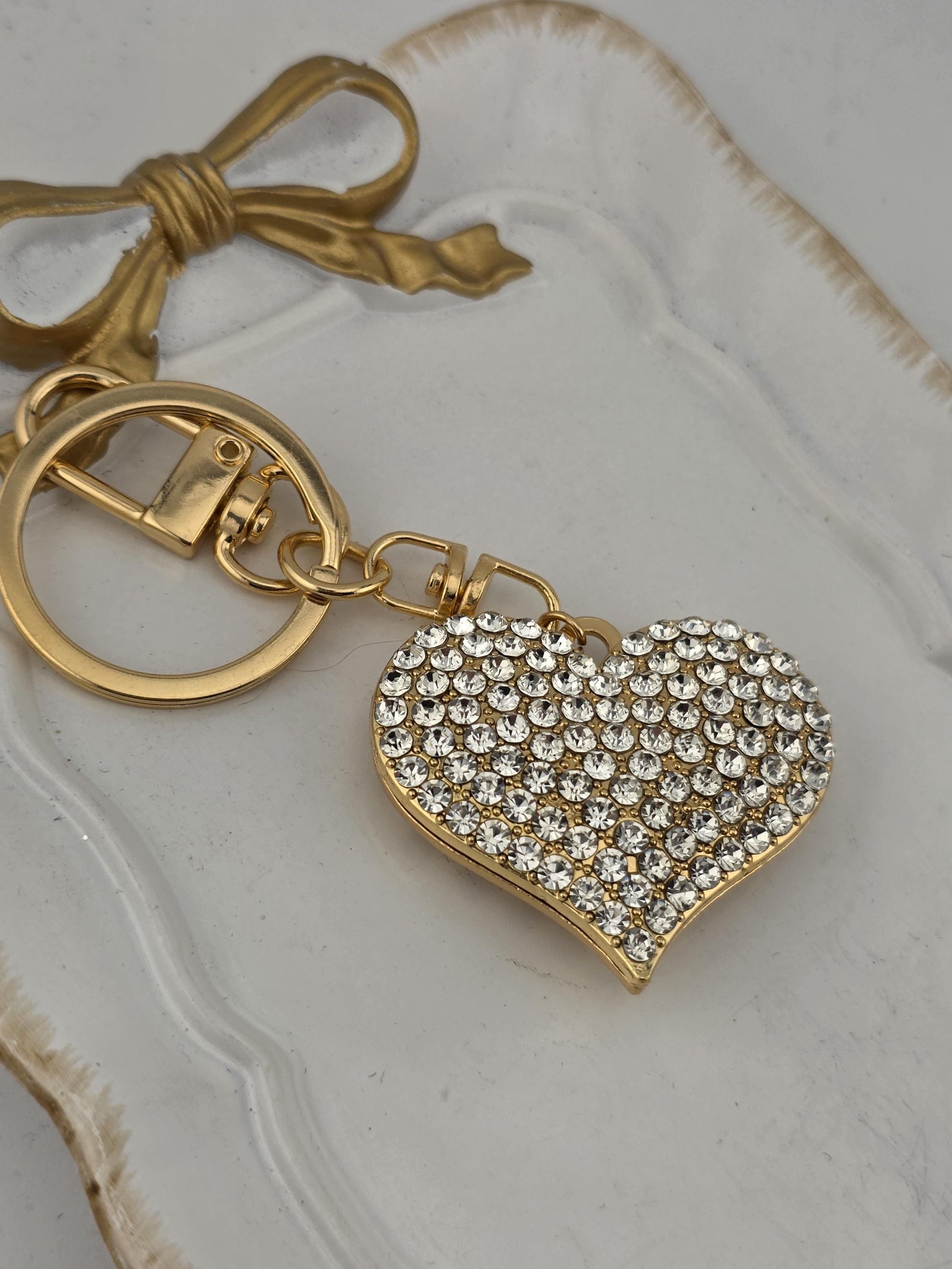 A heart-shaped keychain with a sparkling crystal design is shown on a white surface, accompanied by a gold-colored ribbon bow.