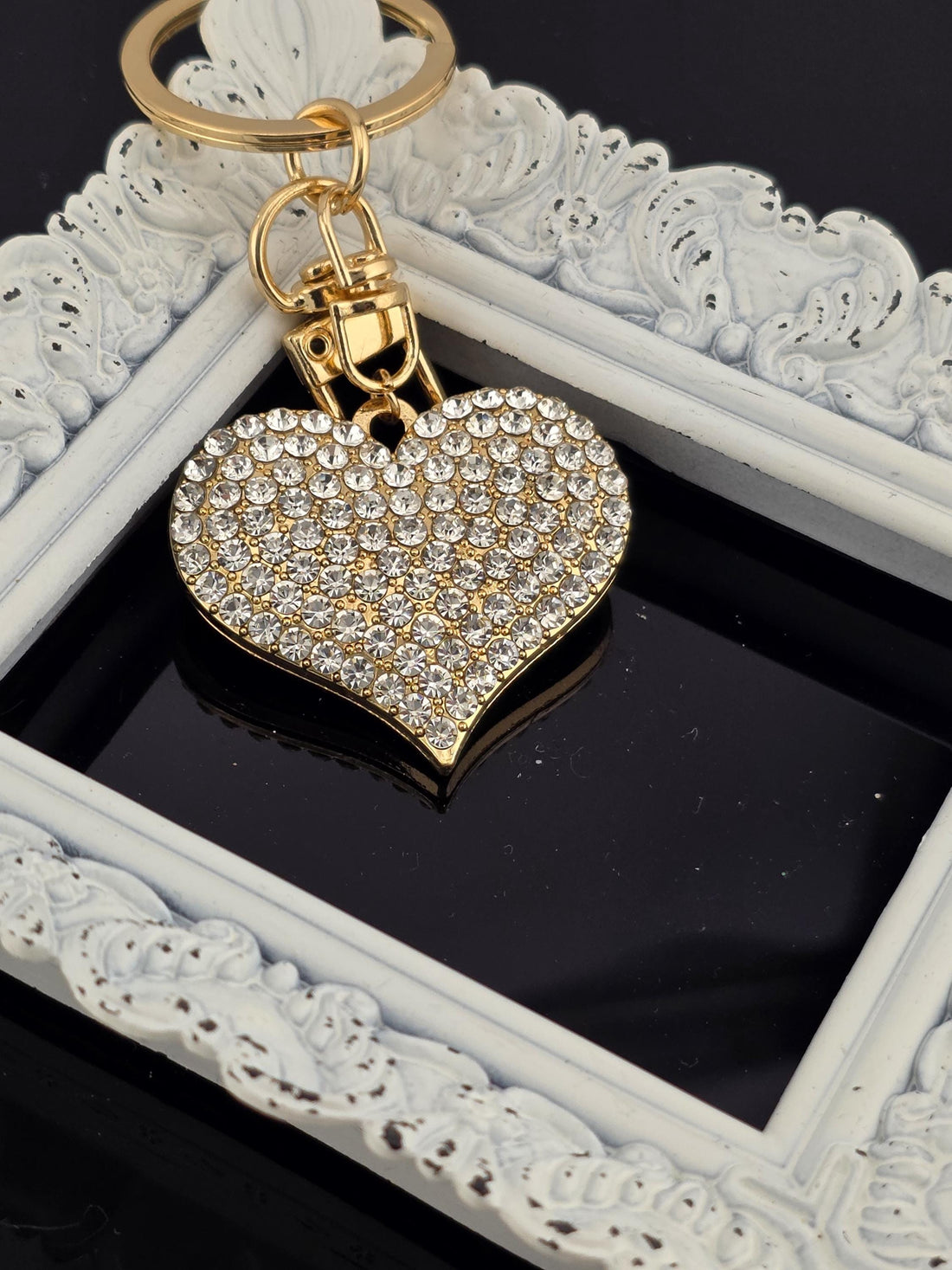A heart-shaped keychain with a sparkling crystal design is shown on a white surface, accompanied by a gold-colored ribbon bow.