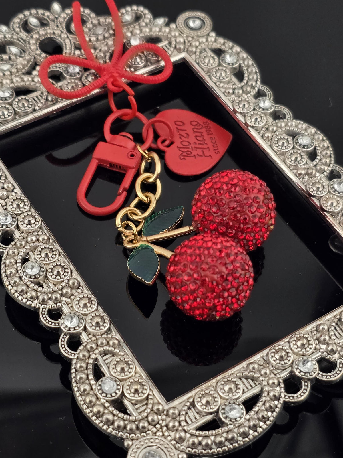 A decorative keychain with a red heart-shaped charm and two red crystal balls, placed on a black surface with a silver frame.