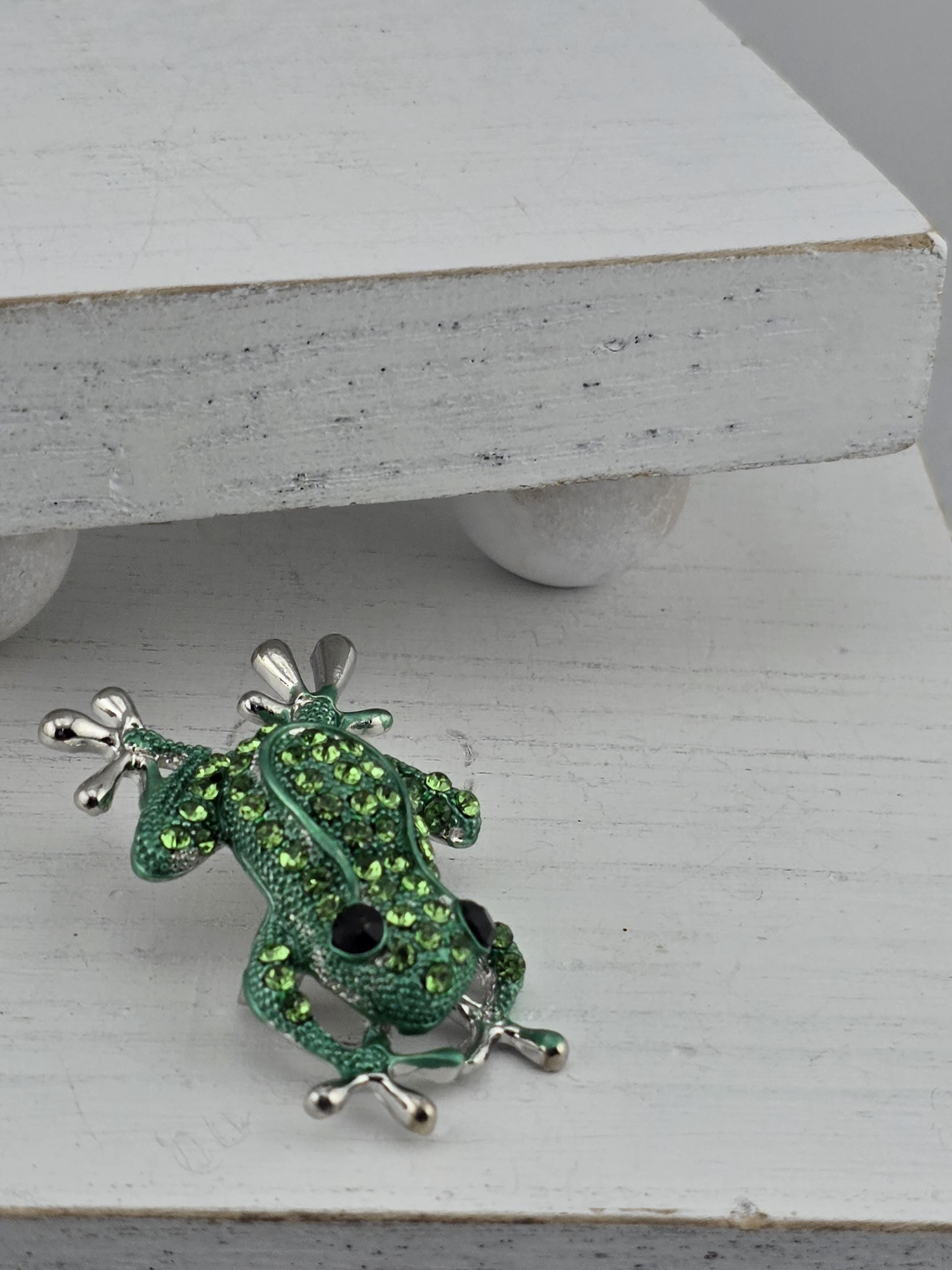 A green frog brooch with rhinestones is displayed on a white wooden surface.