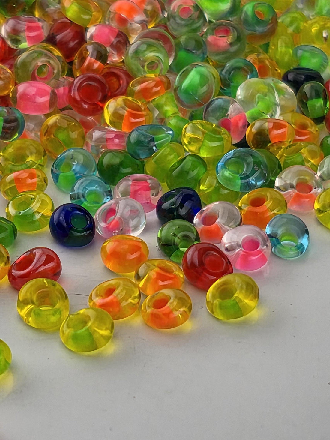 A colorful assortment of multicolored beads, including clear, transparent, and multicolored glass beads, scattered across a white surface.