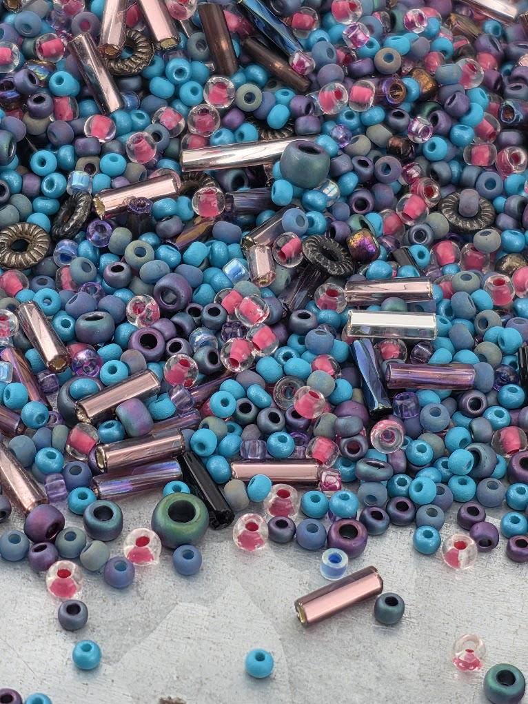 The image is a colorful assortment of various beads and glass pieces, including clear, blue, purple, and pink beads, as well as glass beads and metal pieces.