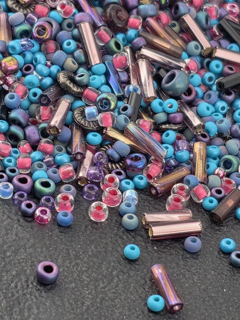 The image is a colorful assortment of various beads and glass pieces, including clear, blue, purple, and pink beads, as well as glass beads and metal pieces.
