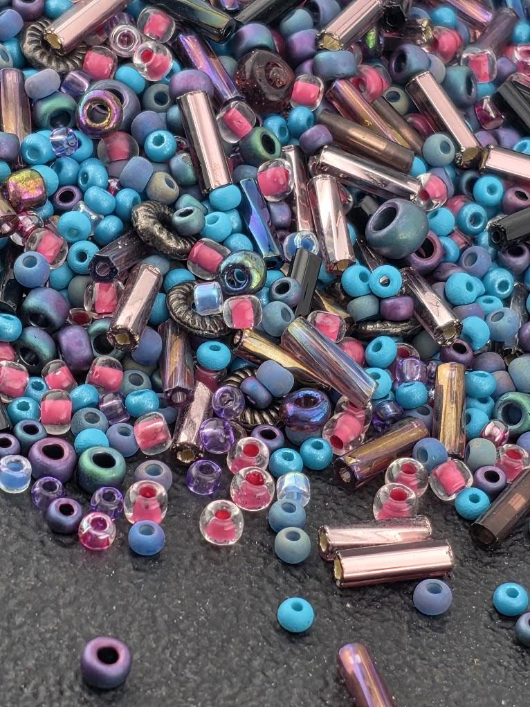 A colorful assortment of glass beads in various shapes and sizes, including clear, blue, purple, and pink beads, scattered on a dark surface.
