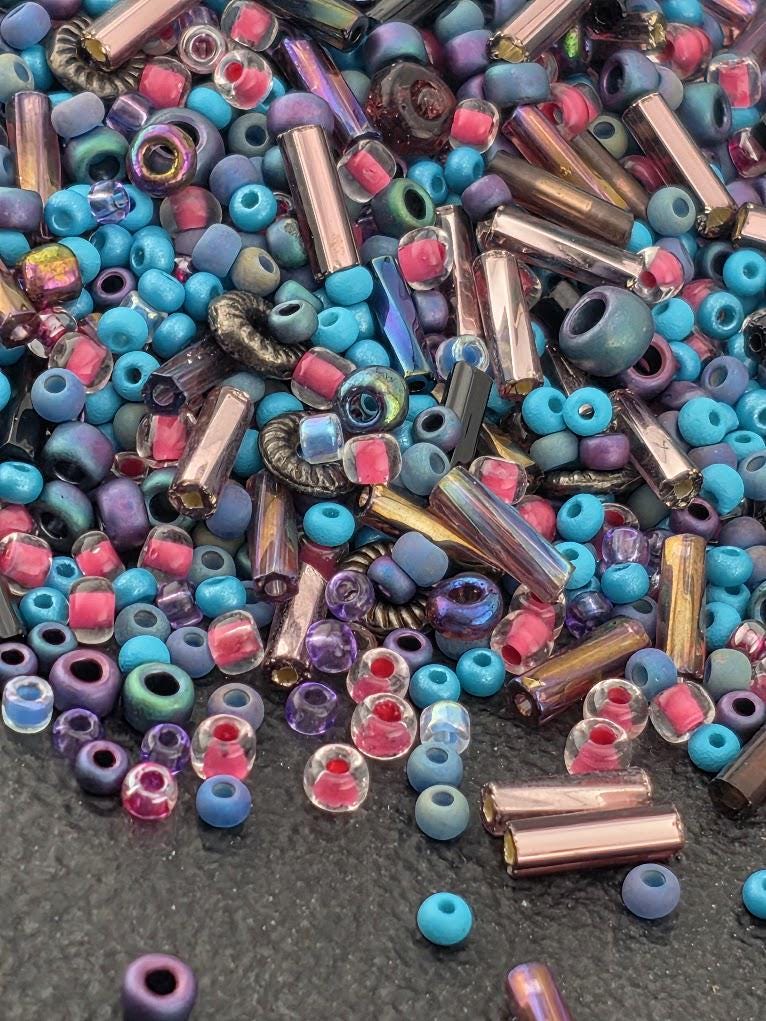 A colorful assortment of glass beads in various shapes and sizes, including clear, blue, purple, and multicolored glass beads, scattered on a dark surface.
