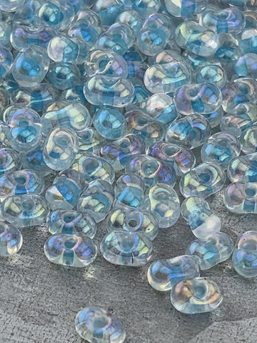 a close-up view of numerous clear, transparent beads scattered on a surface. The beads appear to be made of glass and have a blue-green tint, giving them a unique and iridescent appearance. The image does not contain any text or specific details about the beads, and it does not depict any actions or movements. The focus is on the individual beads and their arrangement on the surface.