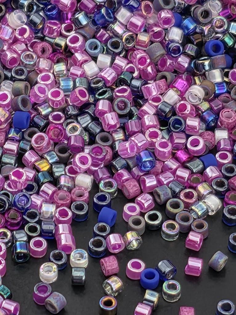 The image is a close-up of a pile of multicolored beads, including various shades of purple, blue, and pink, scattered on a dark surface.