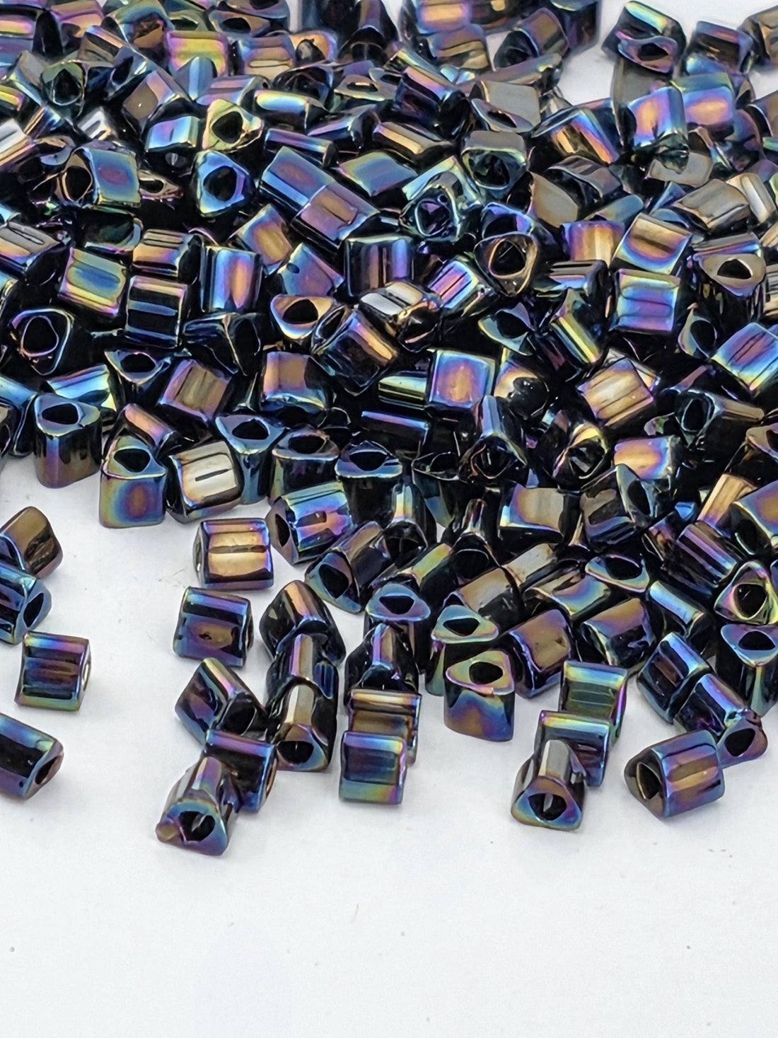 a close-up view of numerous small, shiny metal beads scattered across a white surface. The beads have a metallic, iridescent appearance, reflecting light in various colors. The image does not contain any text or other discernible objects, and the focus is solely on the beads.