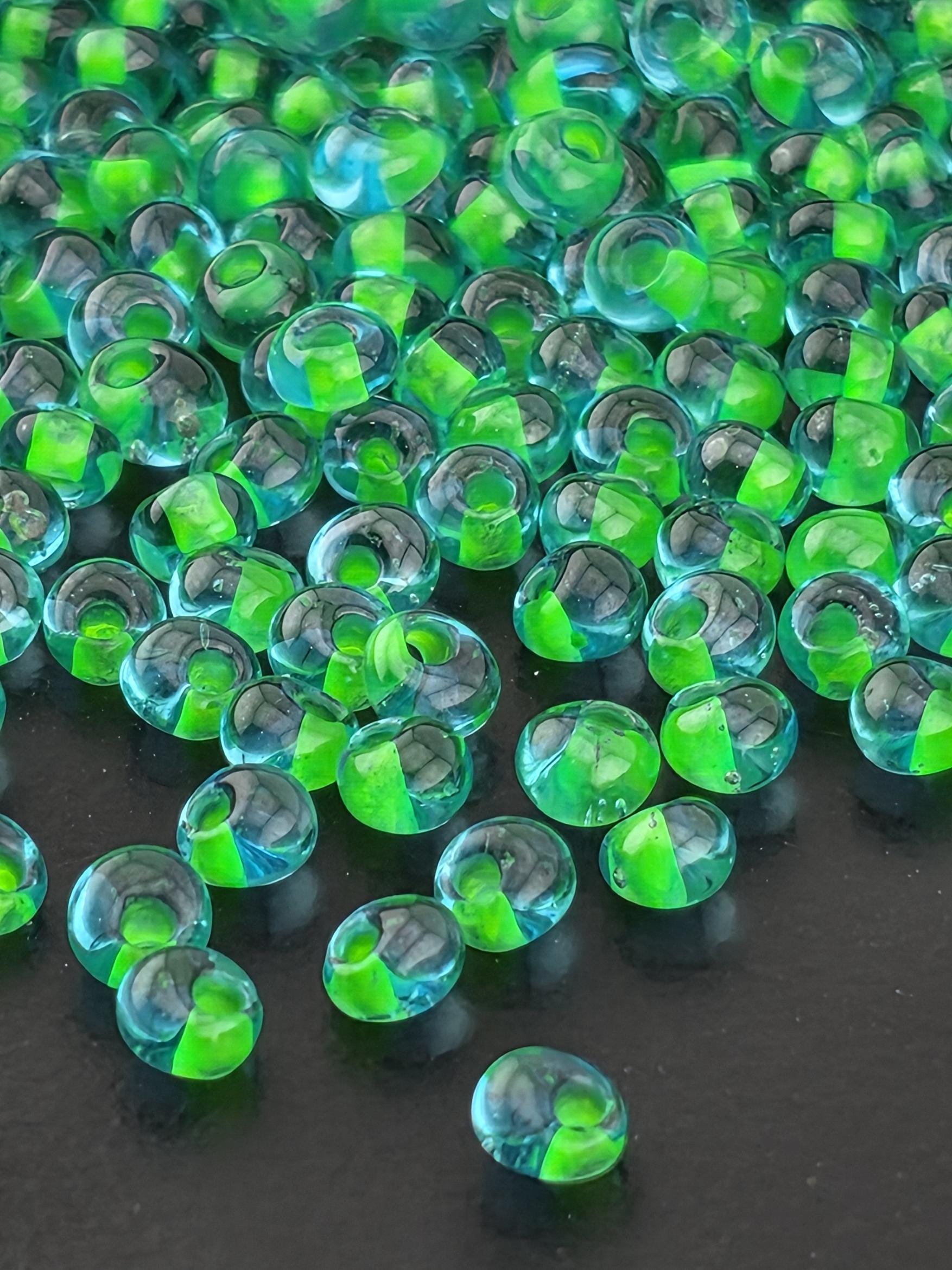a close-up view of numerous small, clear glass beads scattered across a dark background. The beads have a green tint, creating a visually striking contrast against the dark backdrop.