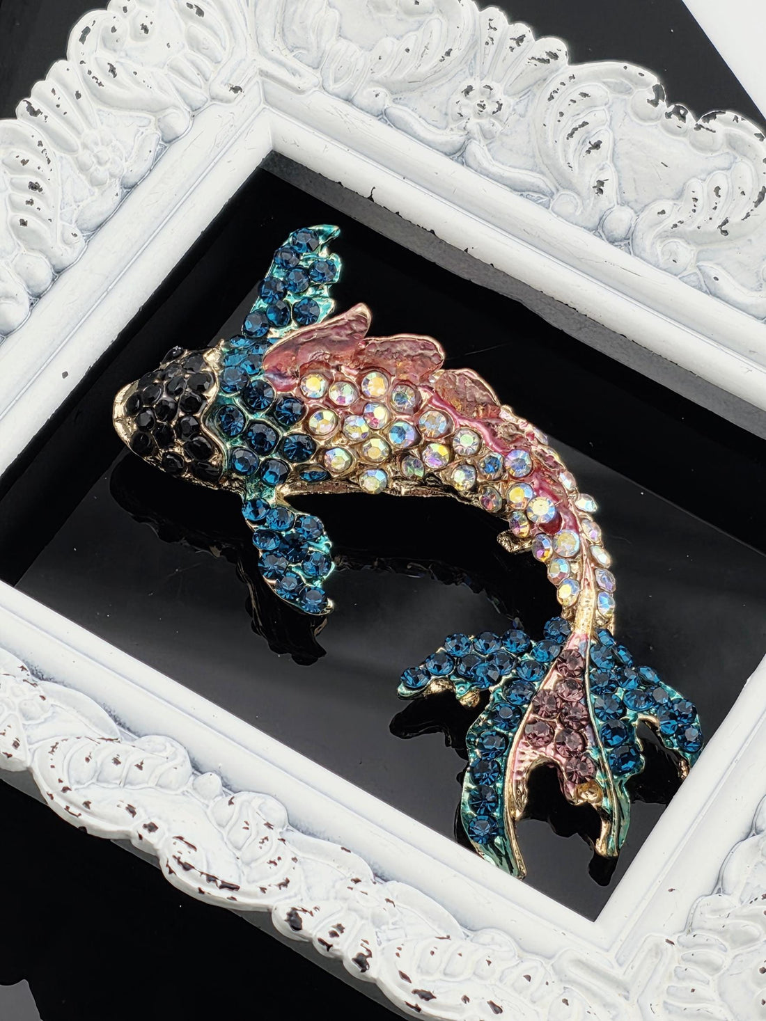 A decorative fish brooch with a colorful, sparkling design is displayed in a white ornate frame.