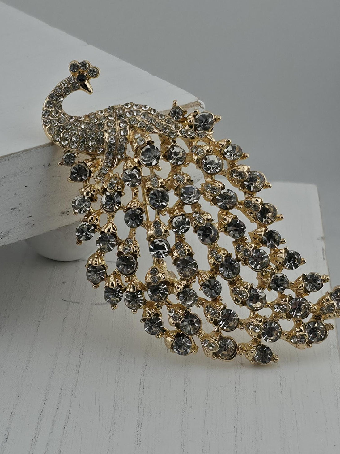 A decorative brooch in the shape of a peacock, adorned with numerous small, clear gemstones in various shades of black and gold.