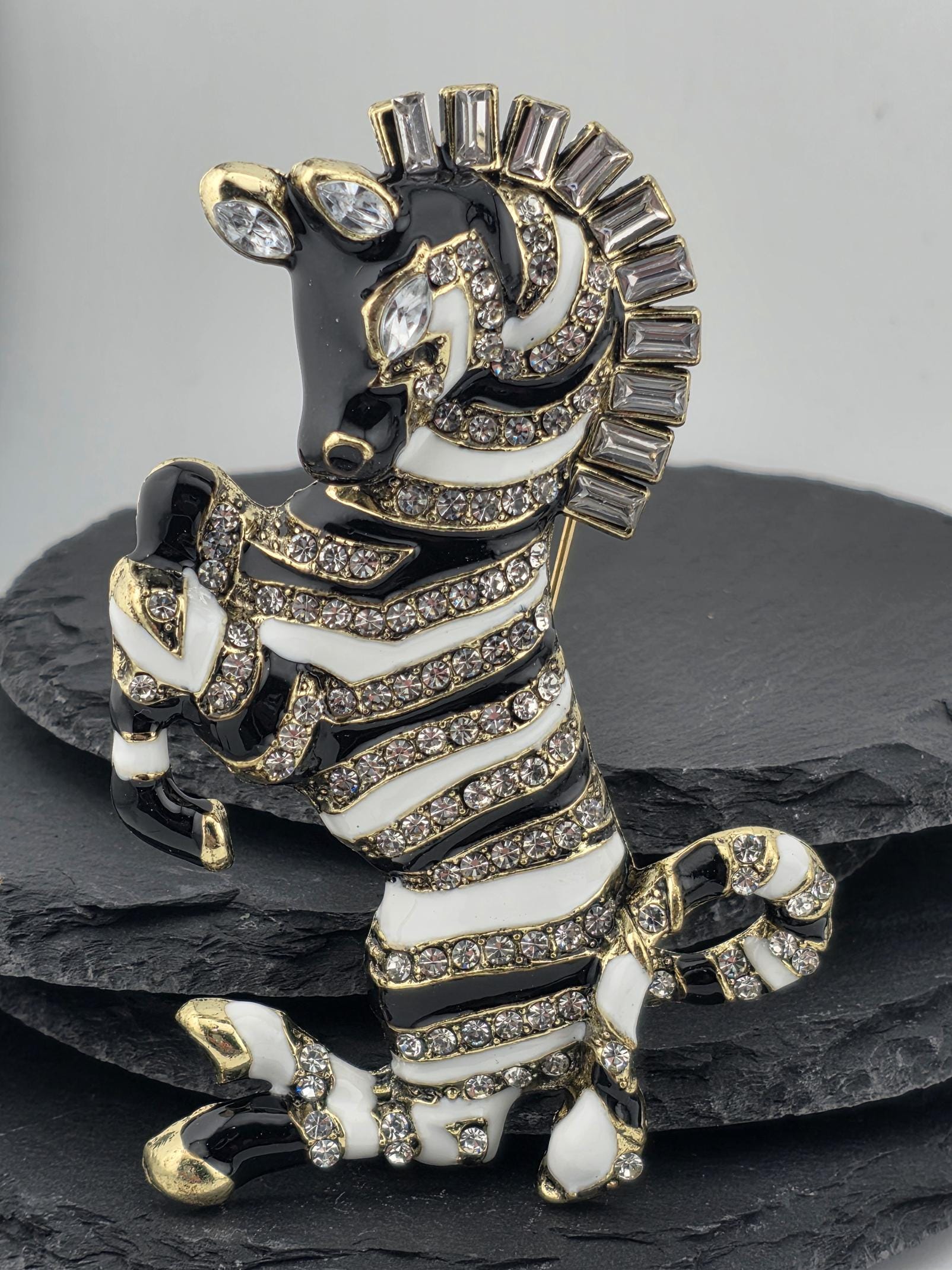 A decorative zebra brooch with a black and white striped pattern, gold accents, and rhinestones or crystals.