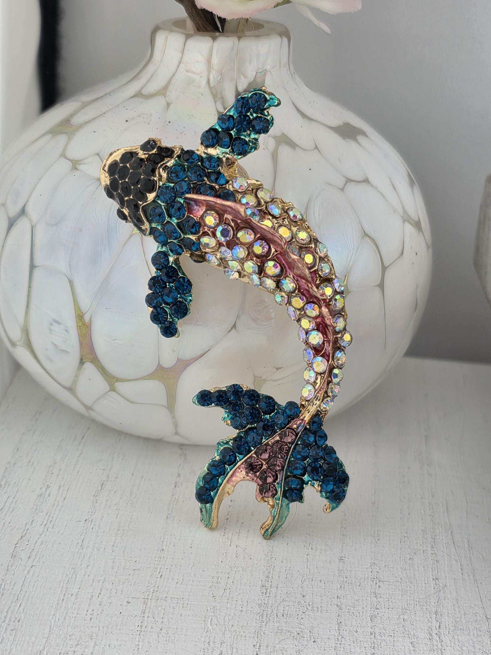 A decorative fish brooch with blue and pink hues, adorned with numerous small rhinestones, is displayed on a white surface.