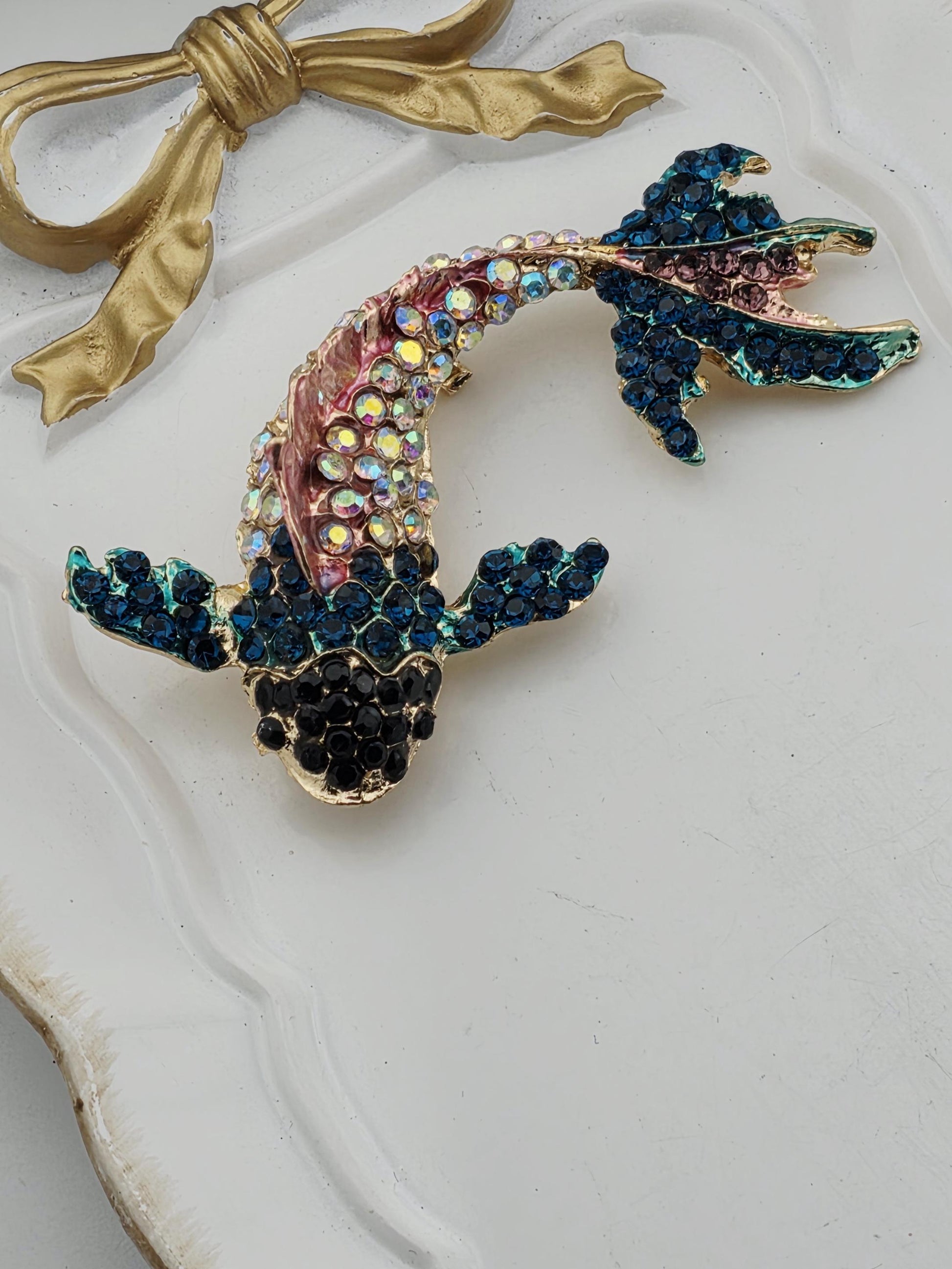 A decorative piece featuring a fish-shaped brooch with blue and green gemstones, placed on a white surface.