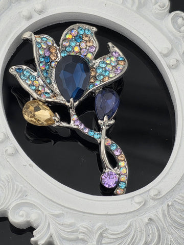 A decorative brooch with a floral design and colorful gemstones, including blue, purple, and yellow stones, set against a black background.