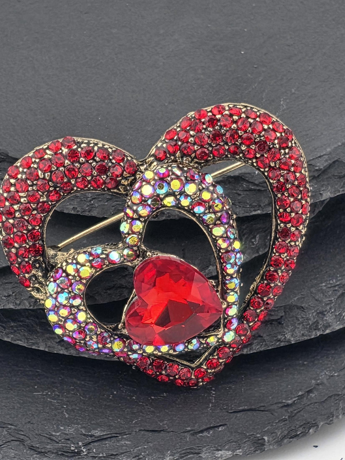 A red heart-shaped brooch adorned with sparkling rhinestones and a large red gemstone is displayed against a black background.