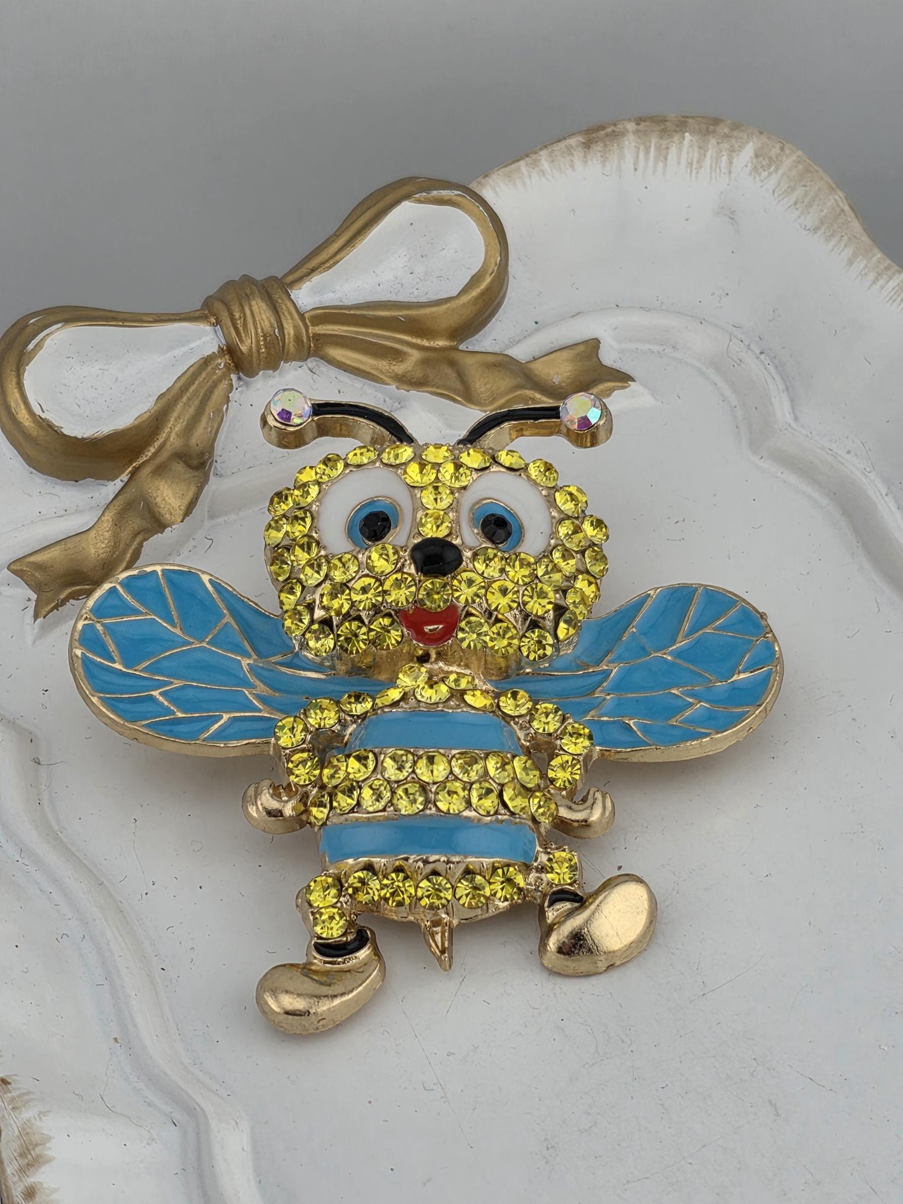 A decorative brooch in the shape of a bee, adorned with yellow and blue gemstones, is displayed on a white surface.