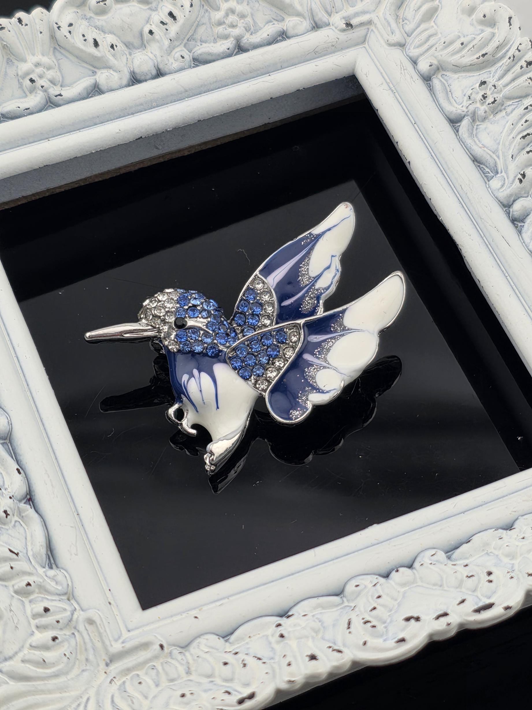 A decorative blue and white butterfly brooch with rhinestones is displayed in a white ornate frame.