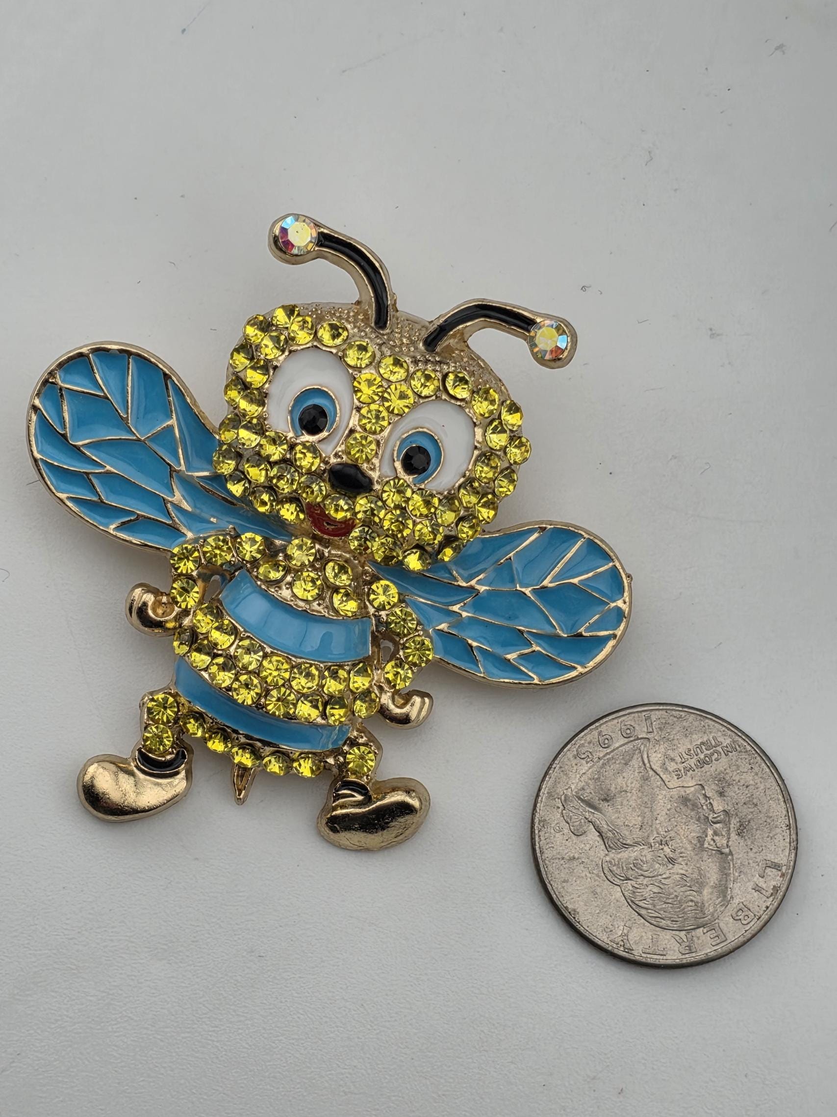 A decorative brooch in the shape of a bee, adorned with blue and yellow gemstones, is placed next to a coin.