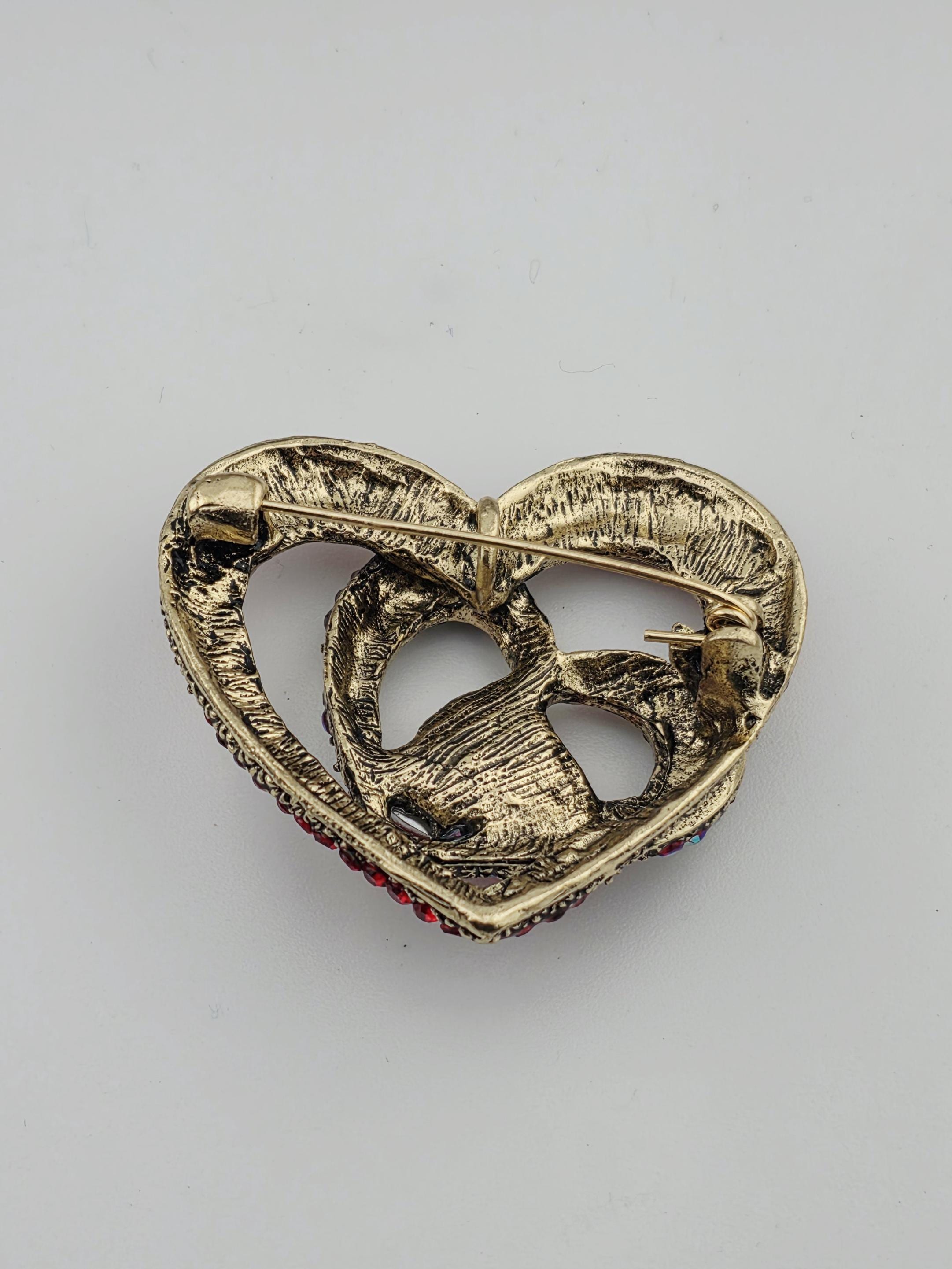 A gold-colored brooch with a heart-shaped design and red gemstones.