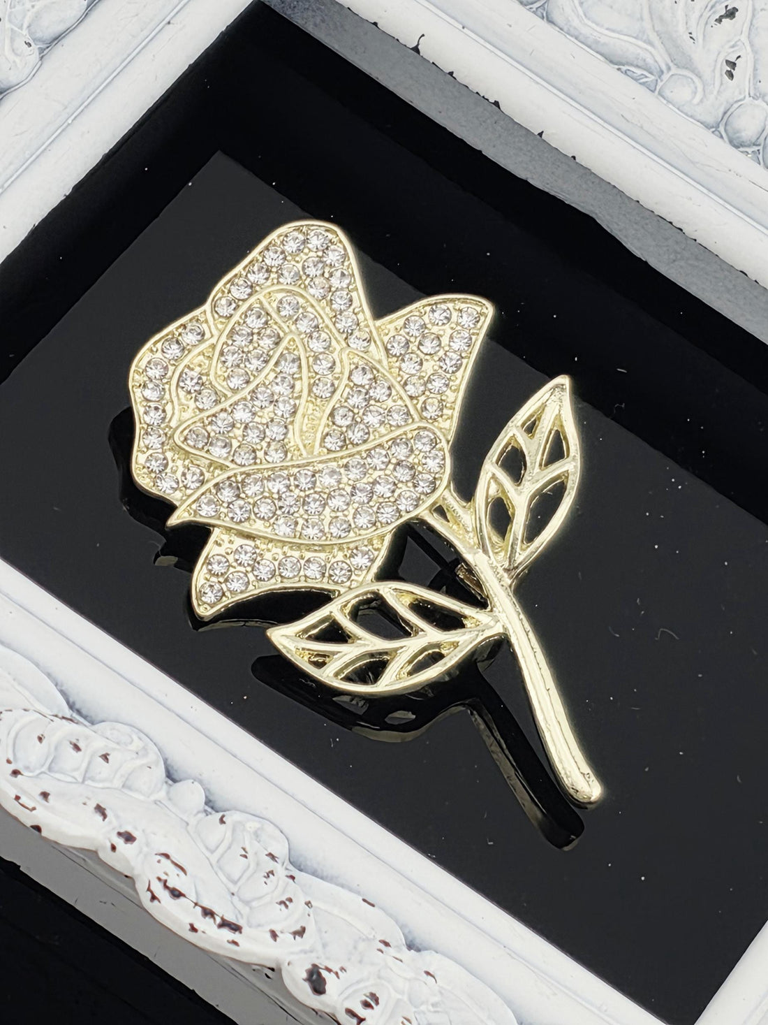 A close-up image of a gold-colored brooch with a diamond-encrusted rose design, placed on a black background.