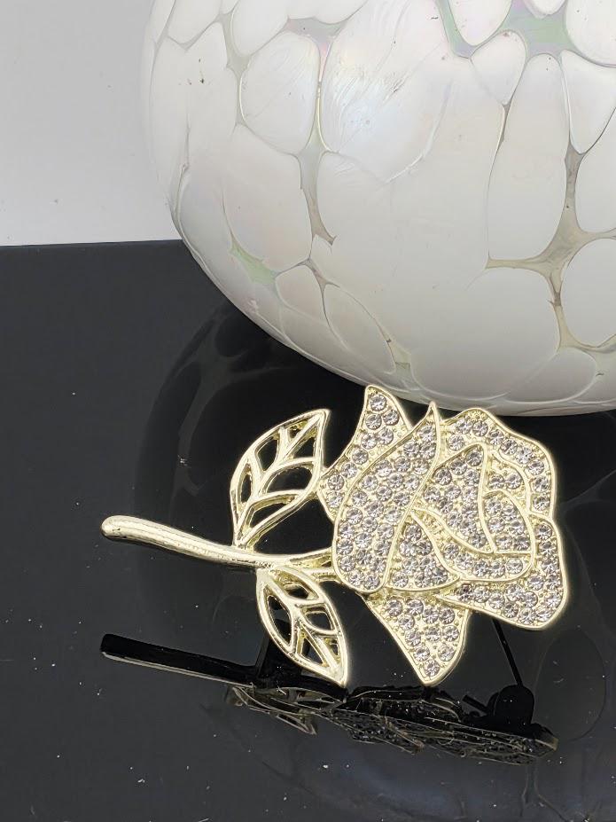 A gold-colored brooch with a diamond-encrusted rose design is displayed on a black surface, with a white vase with a textured surface in the background.