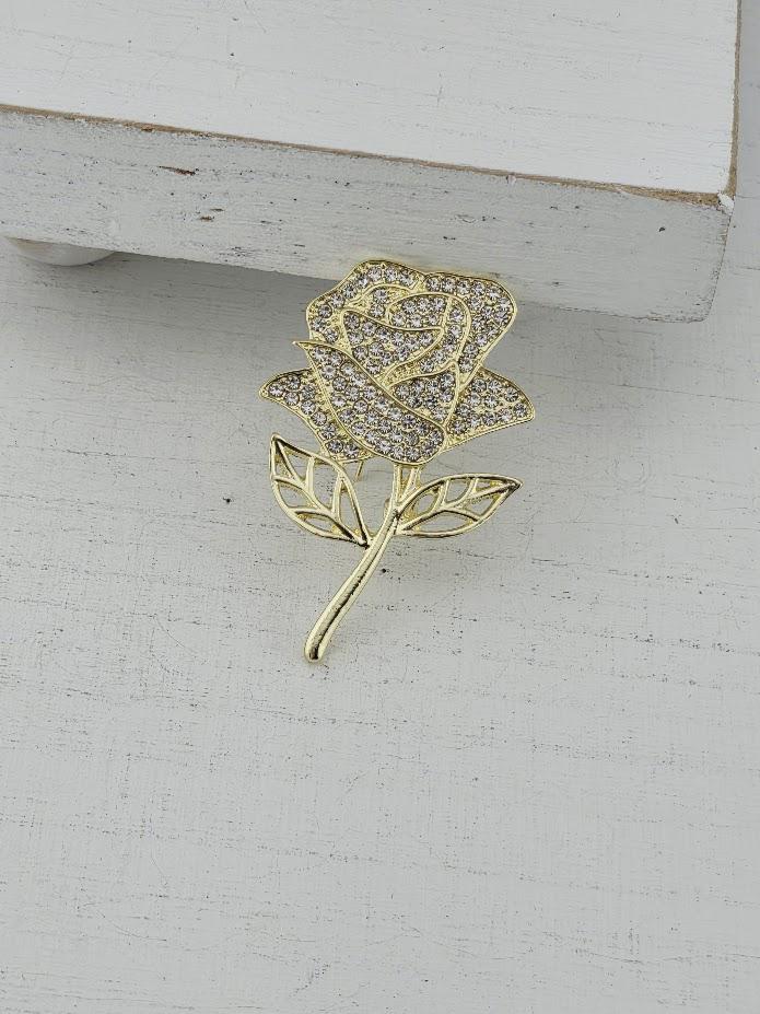 A gold-colored brooch with a diamond-encrusted rose design is displayed on a white surface.