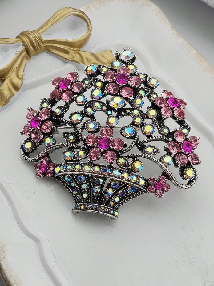 A decorative brooch with a floral design and sparkling gemstones, including pink and purple flowers, is displayed on a white surface.