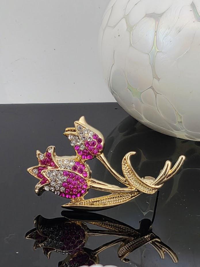 A decorative brooch with a floral design and sparkling gemstones, placed on a reflective surface next to a white vase.