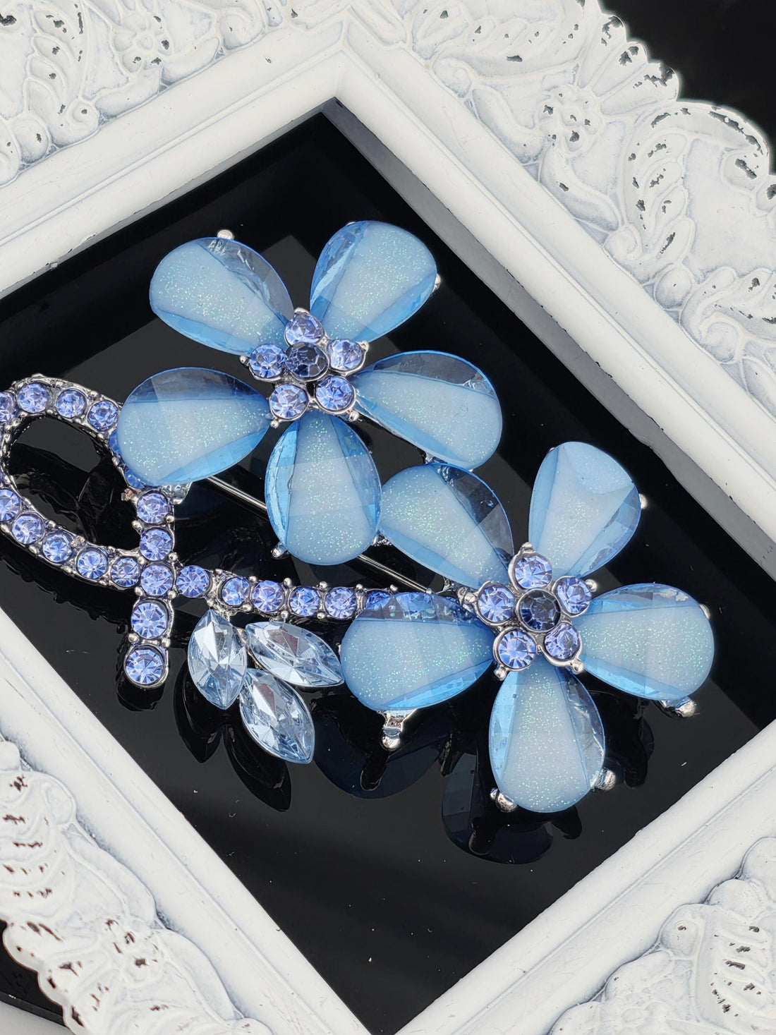 A decorative piece of jewelry featuring blue glass flowers with sparkling gemstones, displayed within a white ornate frame.