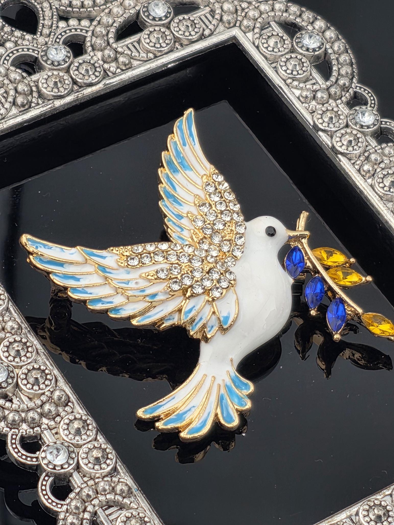A decorative bird brooch with a white body, blue and gold accents, and a gold crown on its head, set against a black background with a silver frame.