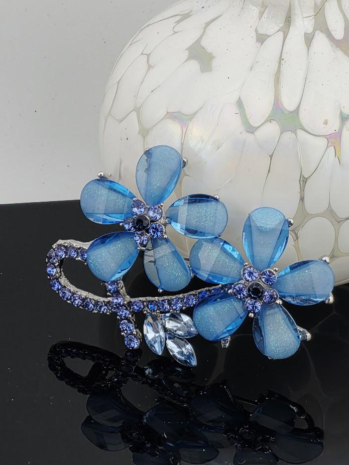 A decorative piece of jewelry featuring blue glass flowers with sparkling gemstones, displayed within a white ornate frame.