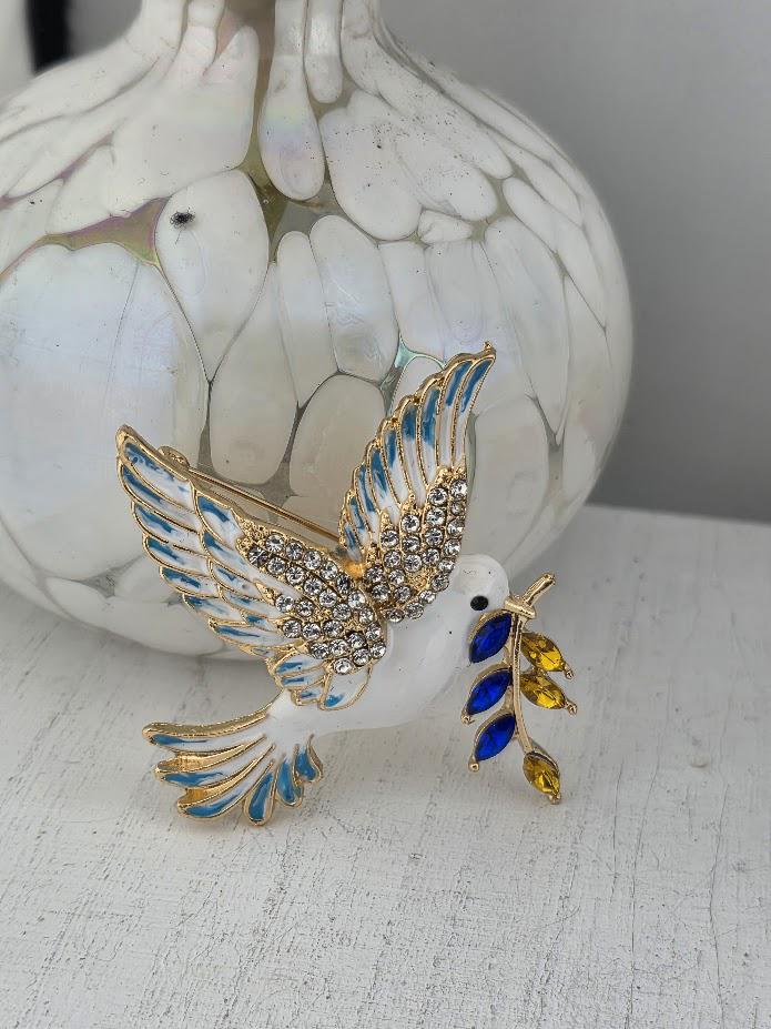 A decorative brooch in the shape of a dove, adorned with blue and yellow gemstones, is displayed on a white surface next to a white vase.