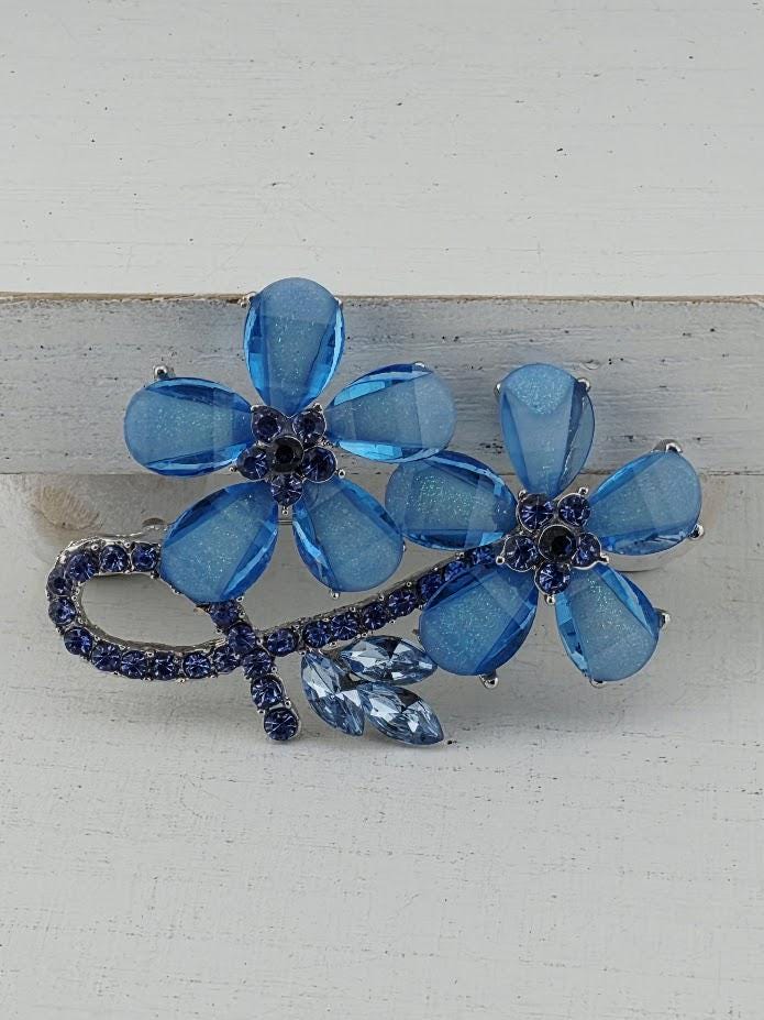 A decorative brooch with blue and clear gemstones, featuring two blue flowers with clear petals and small blue gemstones.