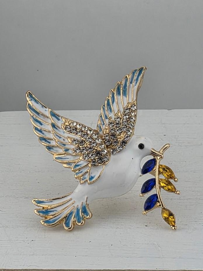 A decorative bird brooch with a white body, blue and gold accents, and a branch with blue and yellow stones.