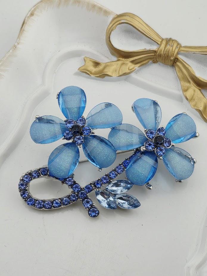 A decorative brooch with blue and clear gemstones, featuring two blue flowers with clear petals and a gold bow.