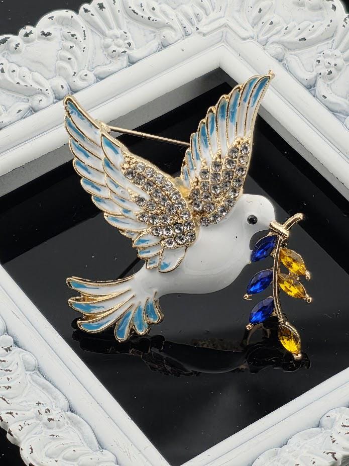 A decorative pin featuring a white bird with blue and yellow accents, holding a branch with colorful stones.