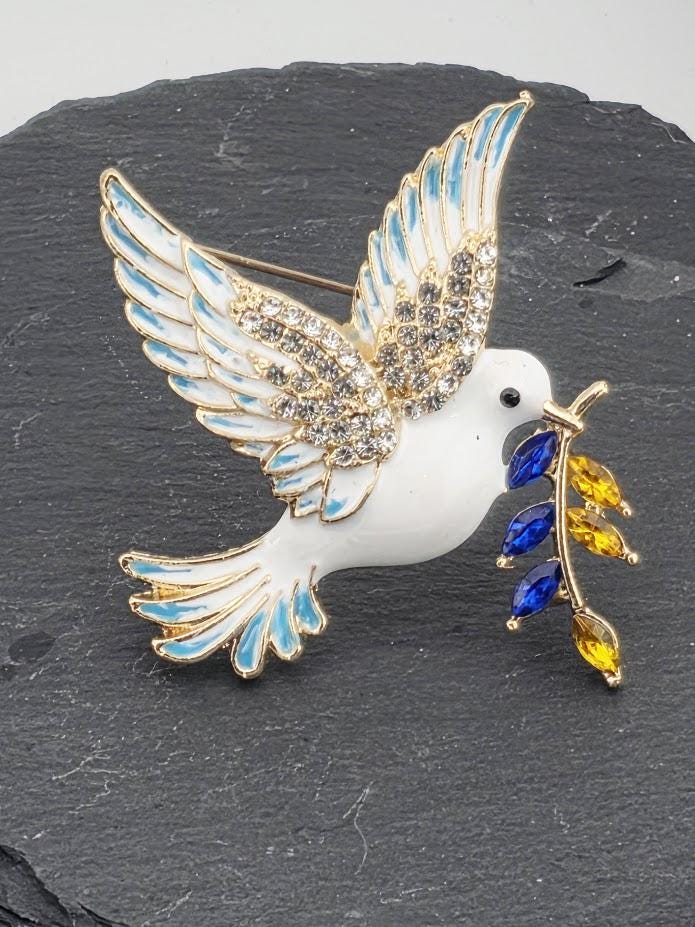 A decorative pin in the shape of a bird, with a white body, blue wings, and a gold beak, holding a leaf with blue and yellow gemstones.
