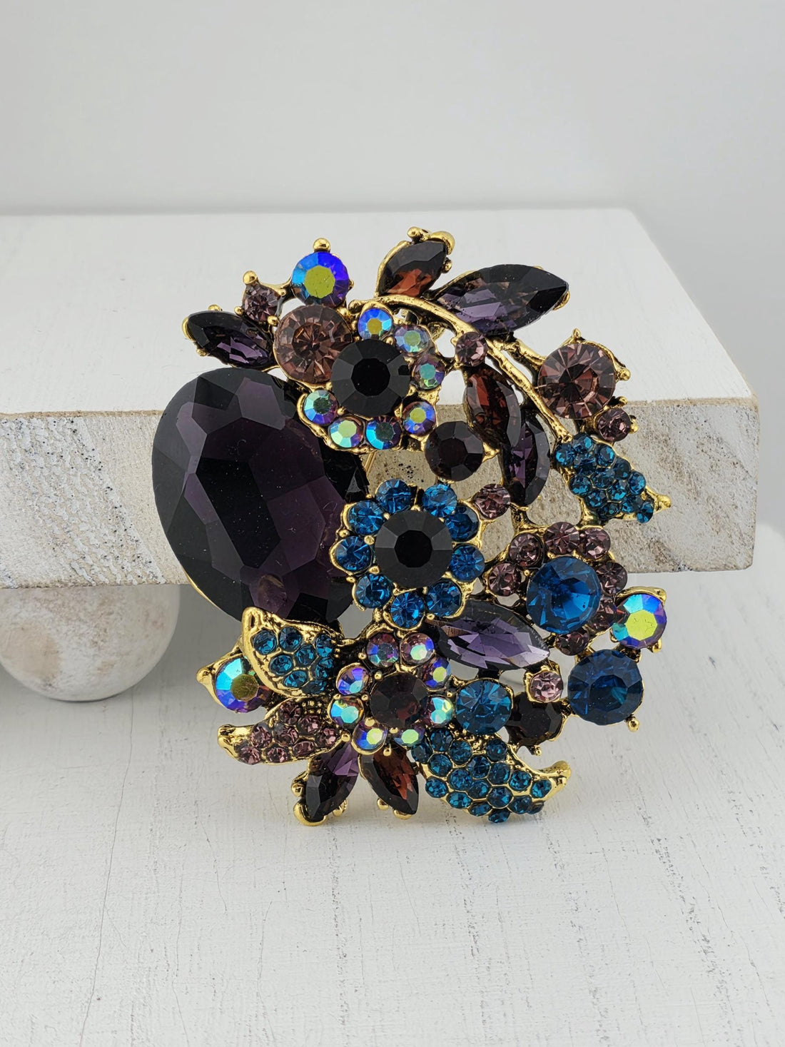 A decorative brooch with a variety of colorful gemstones and floral elements, including purple, blue, and brown stones, as well as some flowers.