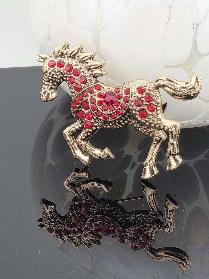A decorative horse brooch with red gemstones on its body and mane, placed on a reflective surface.