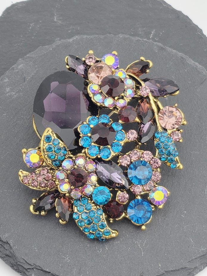 A decorative brooch with a variety of colorful gemstones and floral elements, including purple, blue, and pink stones, as well as some flowers.