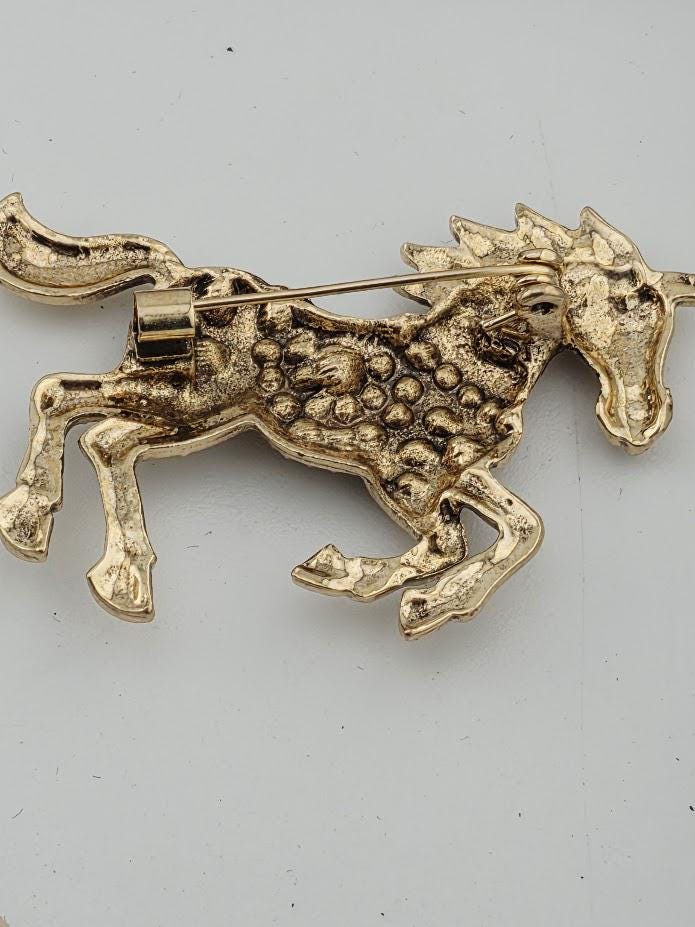 A gold-colored brooch in the shape of a horse, with intricate details and a metallic appearance.