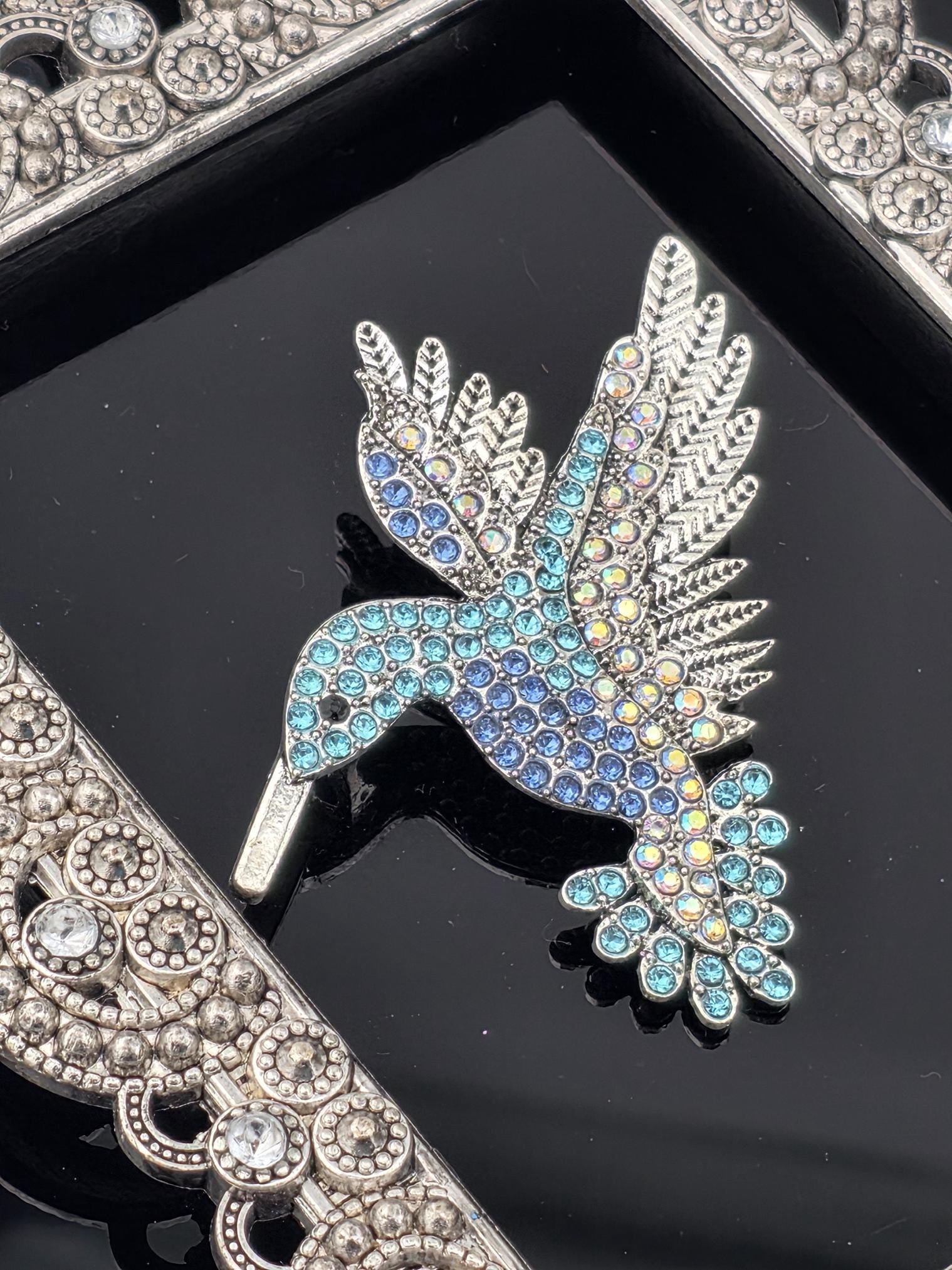 A decorative brooch with a bird-like design, featuring blue and green gemstones, is displayed on a black background.