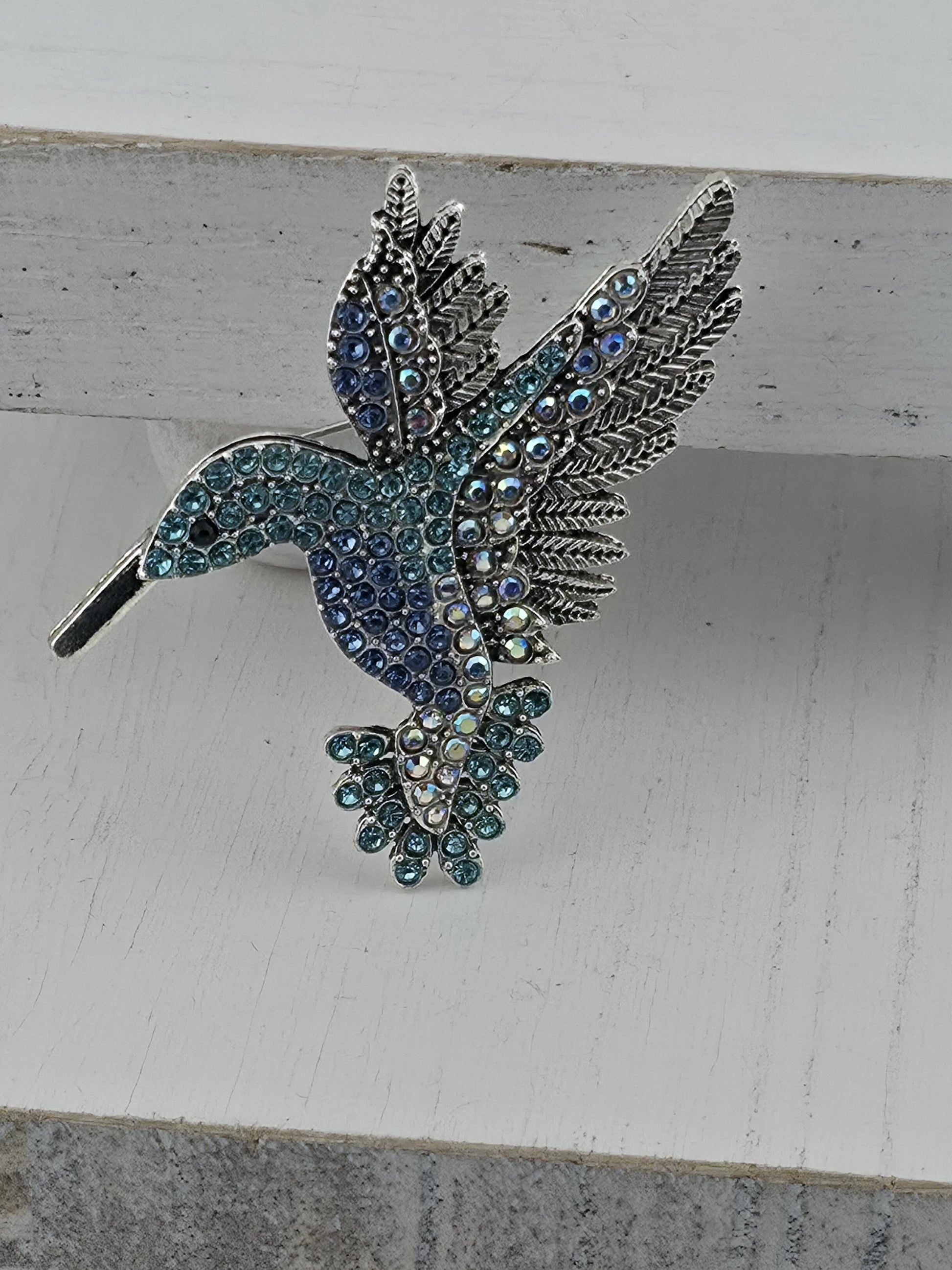 A decorative brooch in the shape of a bird, adorned with blue and green gemstones, is displayed against a white background.