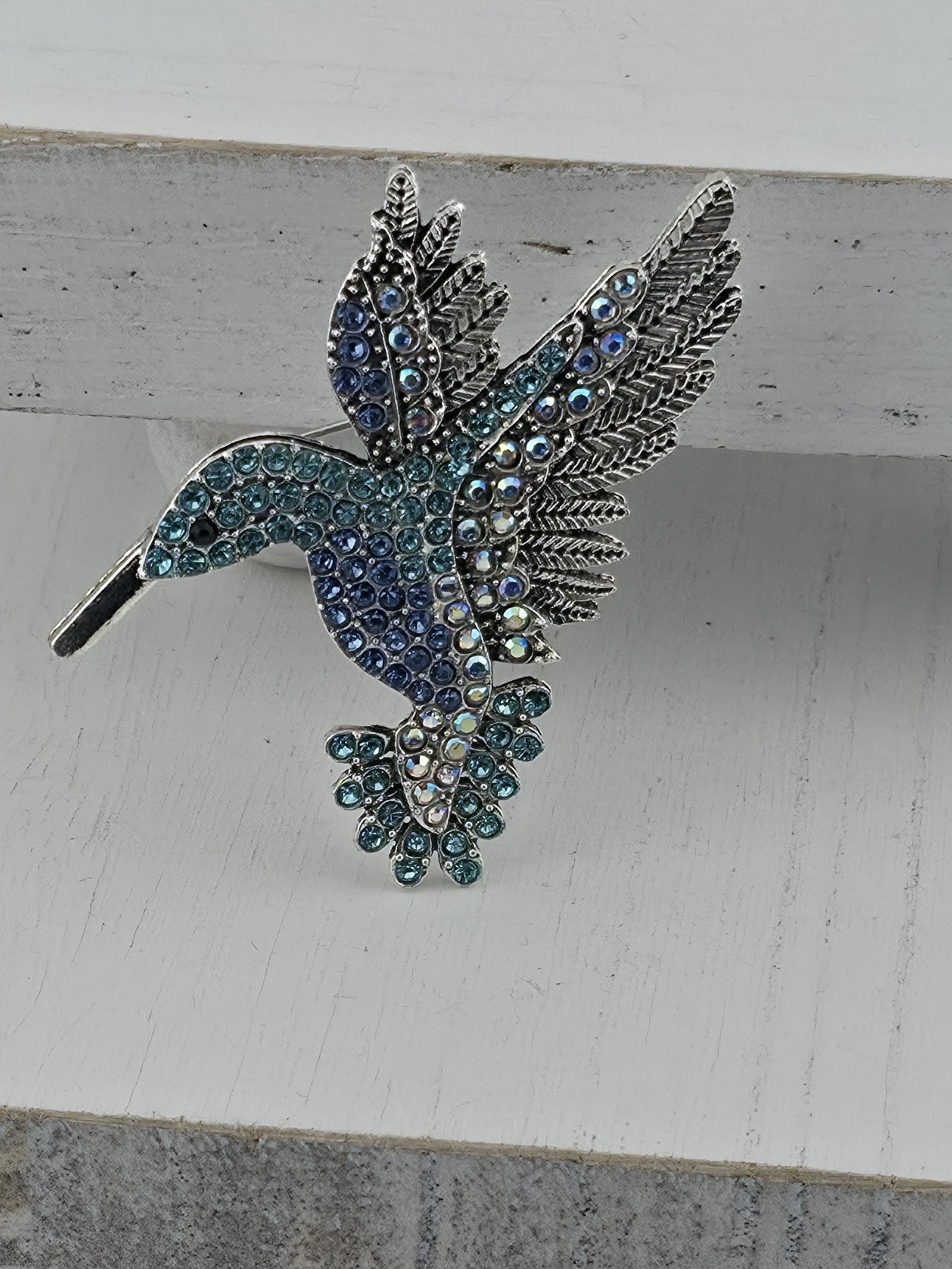 A decorative brooch in the shape of a bird, adorned with blue and green gemstones, is displayed against a white background.