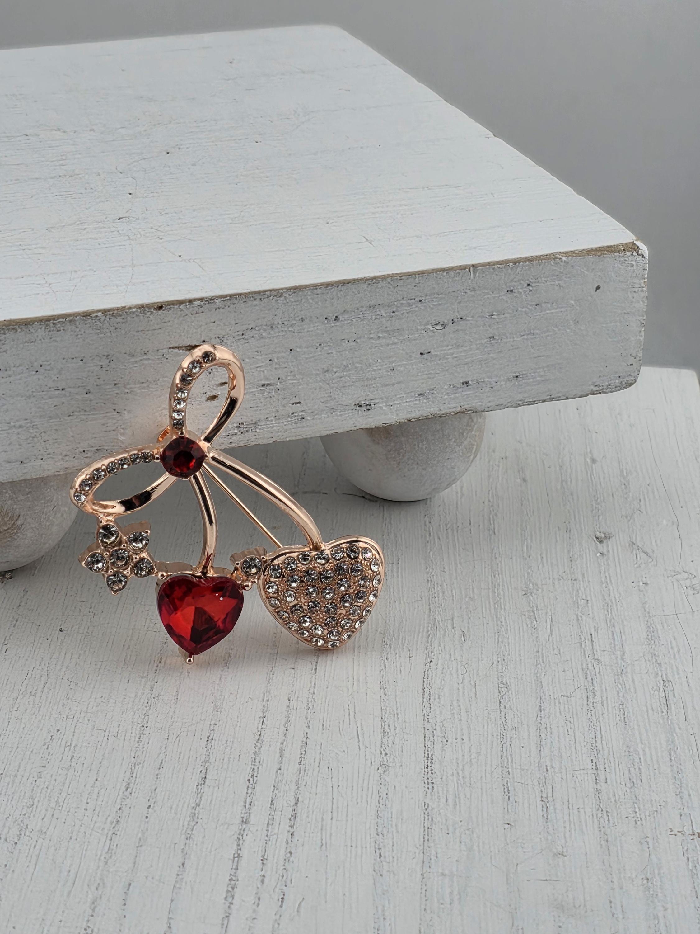 A decorative brooch with a heart-shaped gemstone and a flower-shaped gemstone is displayed on a white wooden surface.